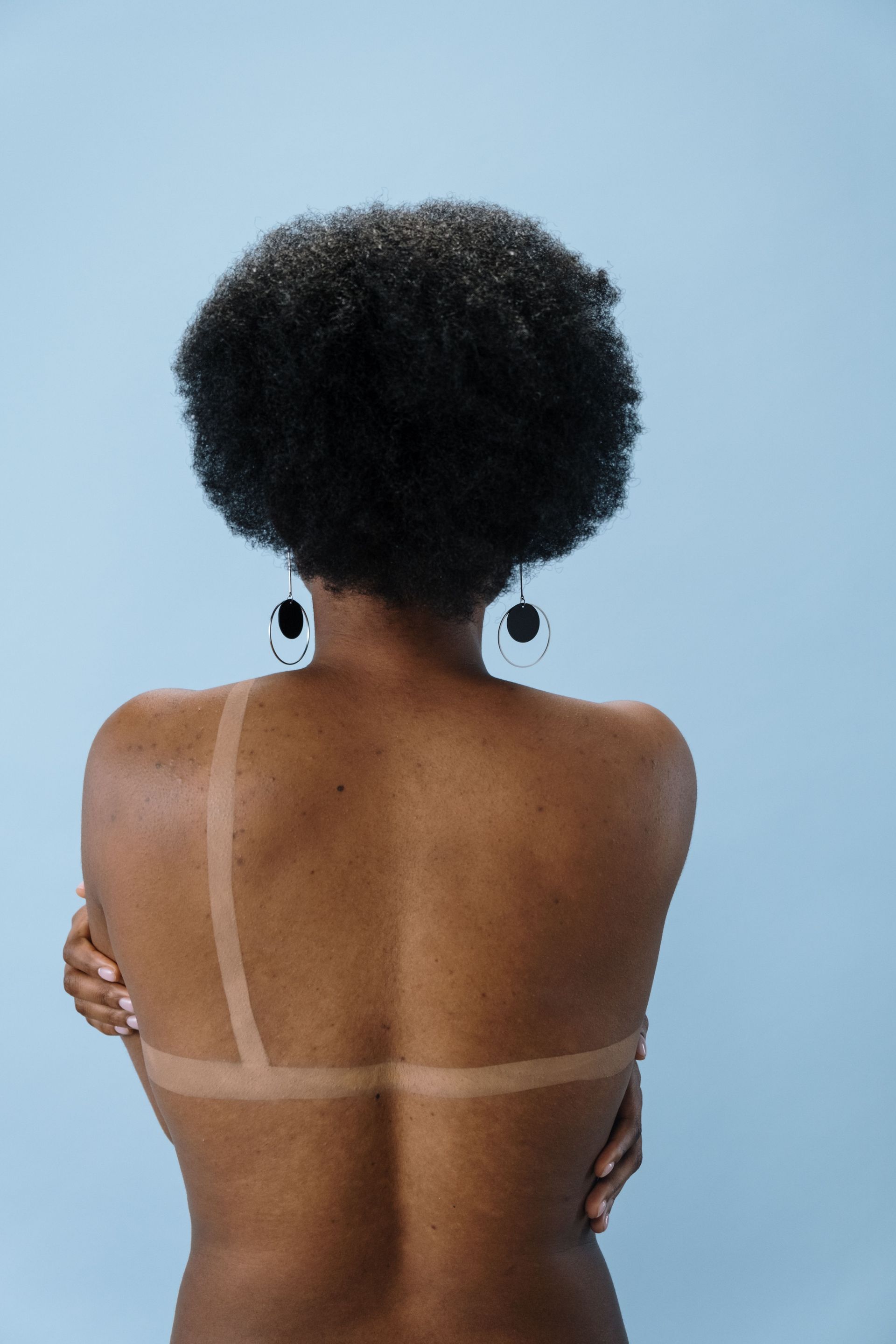 Artistic photo capturing the back view of a shirtless woman with curly hair and hoop earrings against a blue background.